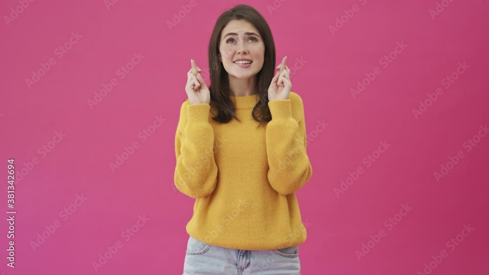 A pleased woman in a yellow sweater is making a wish while crossing her fingers standing isolated over a pink background A pleased woman in a yellow sweater is making a wish while crossing her fingers standing isolated over a pink background