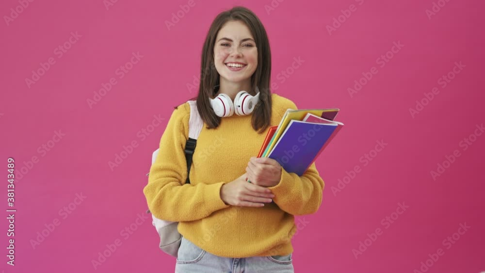 A smiling student woman with headphones is showing thumb-up gesture standing isolated over a pink background A smiling student woman with headphones is showing thumb-up gesture standing isolated over a pink background