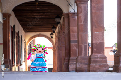 Muñeca de trapo artesanal tradicional con fondo de arco arquitectónico y columnas.