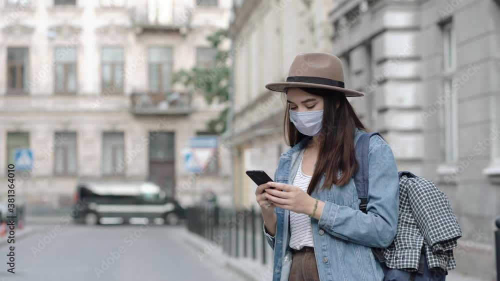 Tourist young woman wearing mask on vacation in old europe city. Portrait of beautiful girl in hat and with backpack. Using smartphone in travel Tourist young woman wearing mask on vacation in old europe city. Portrait of beautiful girl in hat and with backpack. Using smartphone in travel