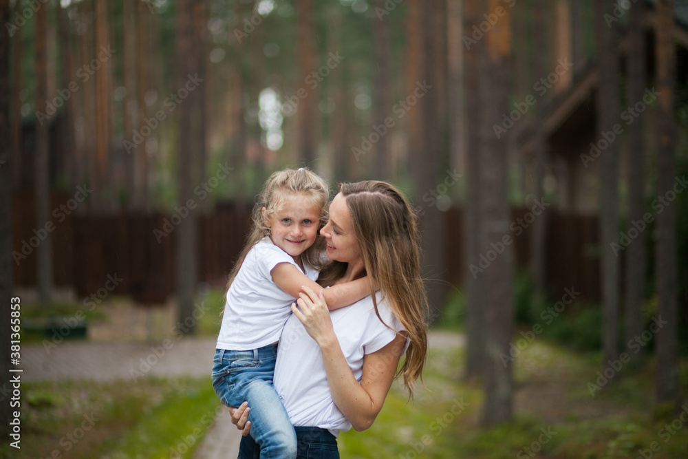 Happy smiling mother and her cute emotional little daughter child in white shirts and denim jeans are hugging and having fun outdoor in nature at countryside. Spending summer time with family.