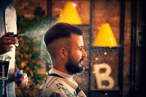Hairdresser using hair spray on client hair at barber shop. Good looking man visiting barber shop. 