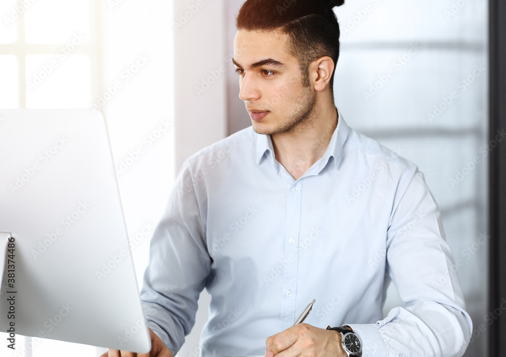 Businessman is working with computer in sunny office. Headshot of arab male entrepreneur at workplace Businessman is working with computer in sunny office. Headshot of arab male entrepreneur at workplace