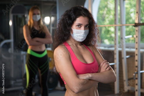 Confident strong woman wearing a protective mask crosses her arms on her chest in the gym ready for a workout. Concept of sports during the pandemic