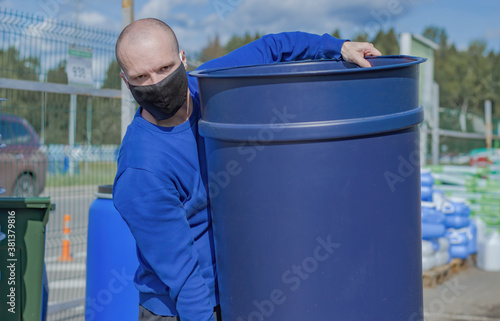 A Male wearing a medical mask carries a blue plastic container. In the background you can see pallets with building materials and an iron fence. photo