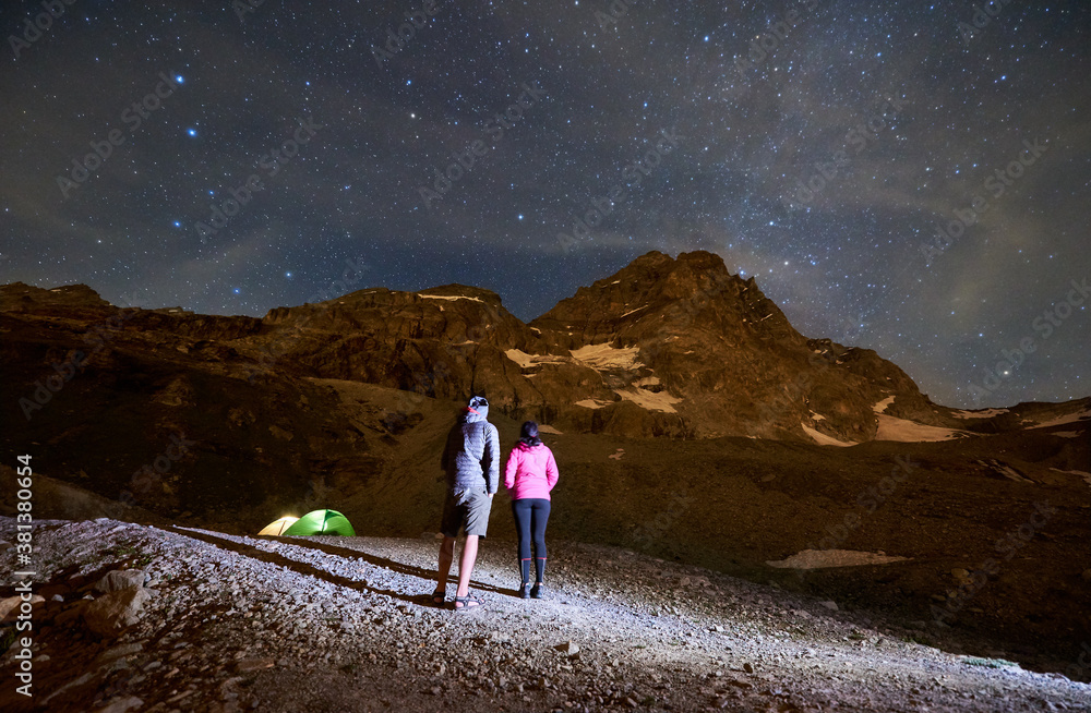 Back view of hikers admiring view of majestic mountains under night sky with starts. Woman and man standing on rocky hill, enjoying night mountain landscape. Concept of travelling, hiking, nighttime.