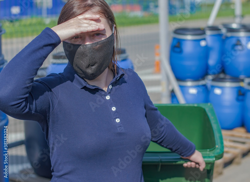 Girl in a protective mask shuts out the sun and looks at the camera. In the girls hand is a green plastic container. An iron fence is visible in the background. photo