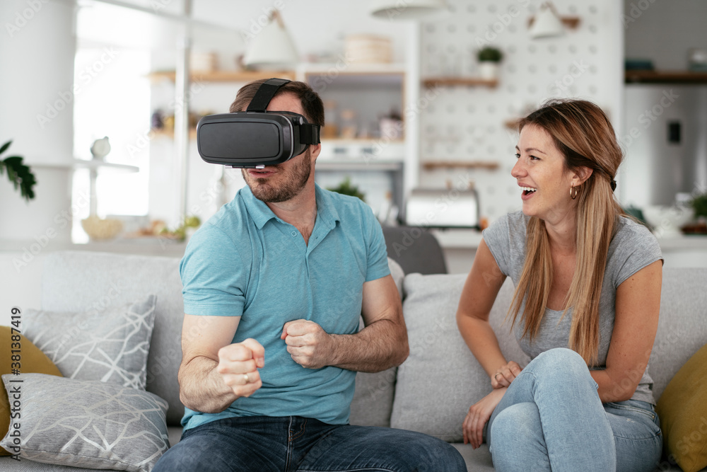 Smiling young man using VR headset at home on couch. Woman and her husband enjoying virtual reality in apartment.. Smiling young man using VR headset at home on couch. Woman and her husband enjoying virtual reality in apartment..