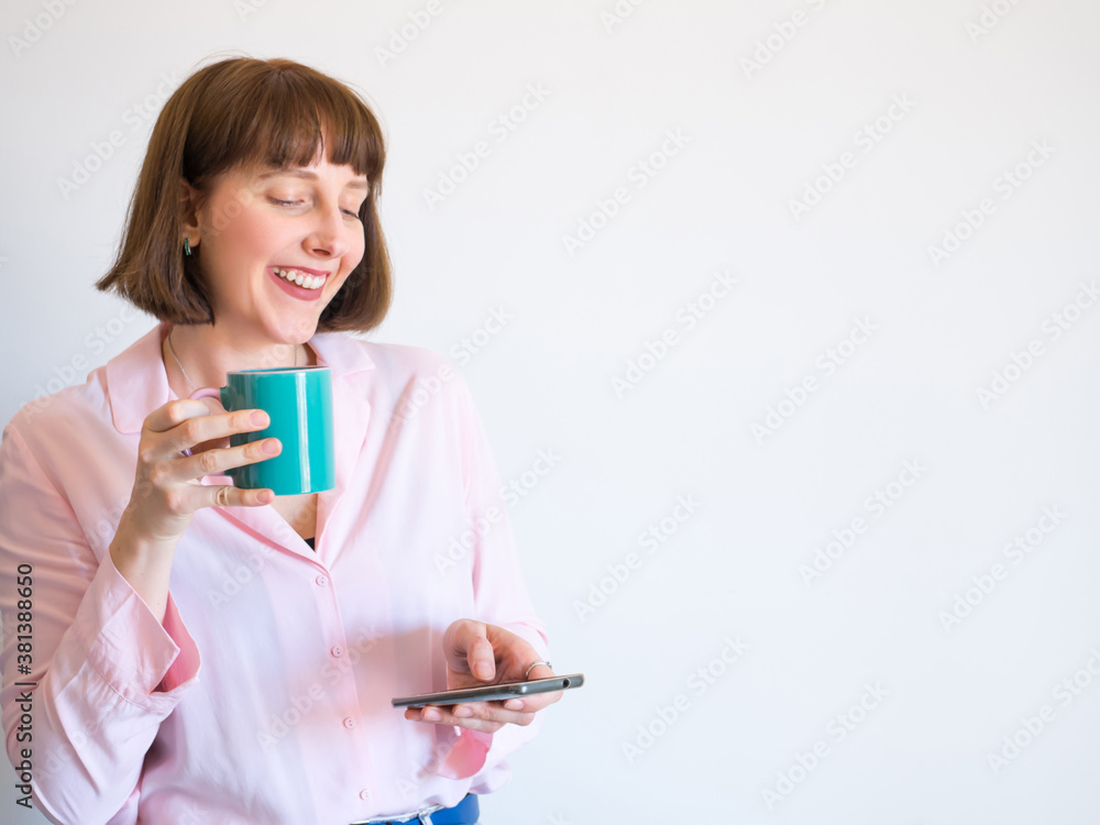 Smiling young woman in jeans and pink shirt drinking coffee and using smartphone on white wall background. Smiling young woman in jeans and pink shirt drinking coffee and using smartphone on white wall background.