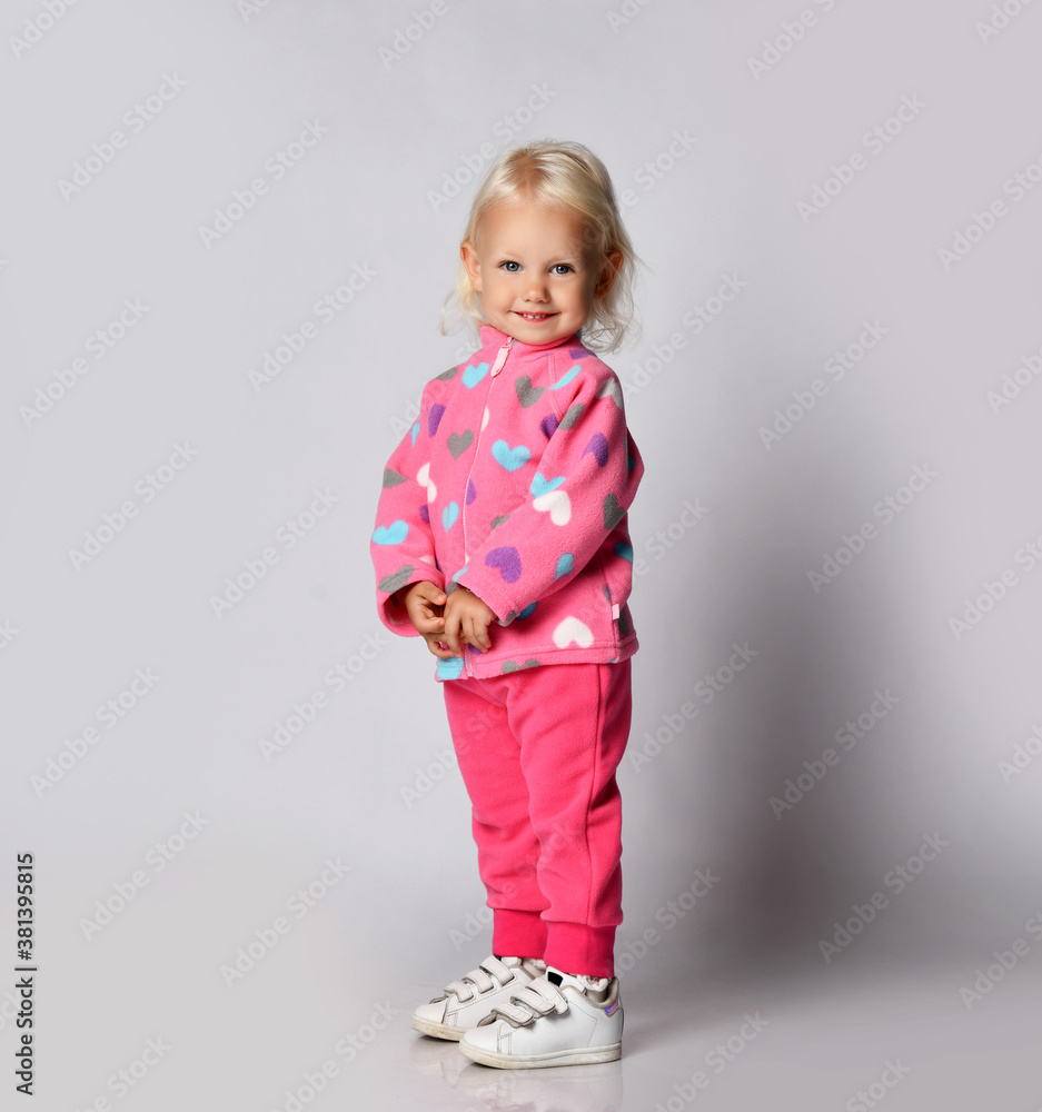Cute blonde smiling baby kid girl in pink warm fleece clothing with heart print pattern stands side looking at camera Cute blonde smiling baby kid girl in pink warm fleece clothing with heart print pattern stands side looking at camera