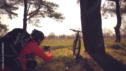 Young caucasian woman athlete tourist cyclist makes photo on phone, photographs his bike standing near tree on nature field sunny day. Girl in helmet and sportswear resting break mountain bike photo