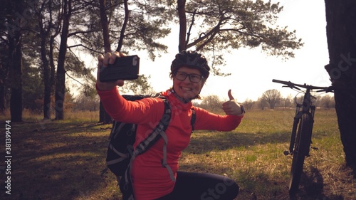 Young caucasian woman athlete tourist cyclist uses hand smart phone photo of herself selfie sitting near tree in coniferous forest outside the city. Sportswoman taking selfie with her mountain bike photo