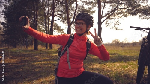 Young caucasian woman athlete tourist cyclist uses hand smart phone photo of herself selfie sitting near tree in coniferous forest outside the city. Sportswoman taking selfie with her mountain bike photo