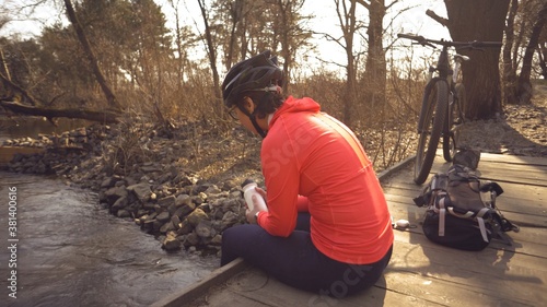 Caucasian woman sportsman in bright sports shoes helmet rests on the bridge cross the river. Cyclist in bright sportswear with a flask a plastic bottle in his hand. Rest stop break drink water thirst