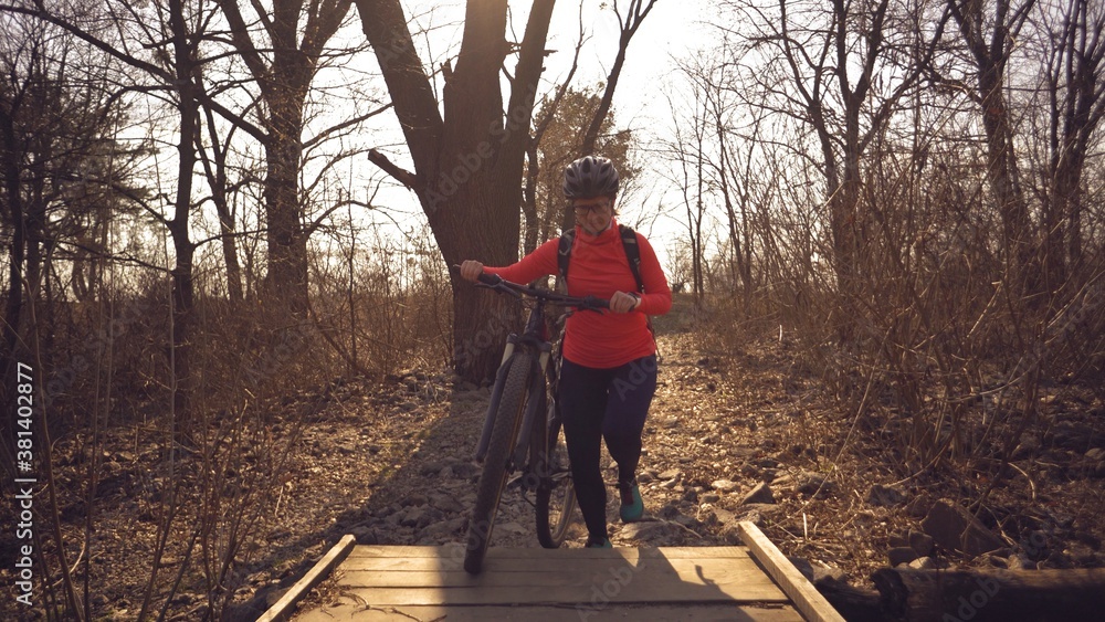 Athlete bicyclist Caucasian woman in sportswear and helmet crosses on foot, leads a mountain bike in her hands across a rural narrow wooden bridge across a small river in the forest