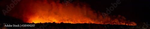 Horizontal panoramic view of a wildfire in a natural setting at night with lots of smoke. Ecological disaster concept