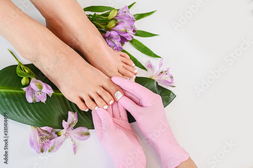 Beautiful perfect skin female legs feet top view with tropical flowers and green palm leaf. Hands of beautician in pink gloves. Nail polish, care and clean, spa pedicure treatment in white. Copy space