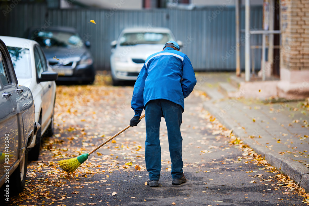 Worker cleans yard area from fallen autumn leaves with broom tool. Man sweep and cleaning road from fallen yellow leaves. Janitor sweep leaves from road. Municipal worker with broomstick