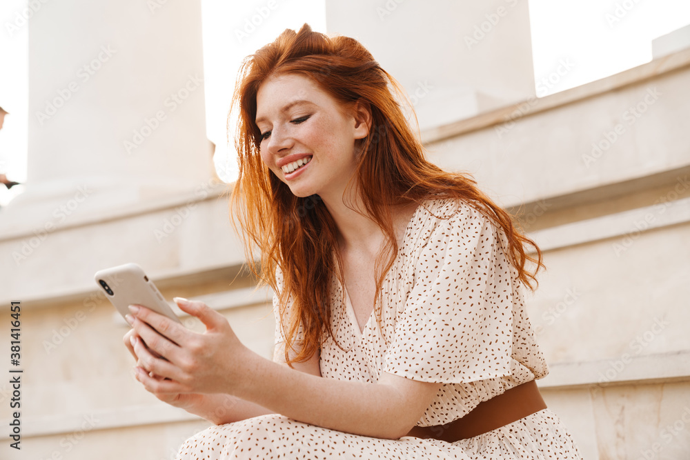 Image of cheerful ginger girl using mobile phone while sitting on stairs Image of cheerful ginger girl using mobile phone while sitting on stairs