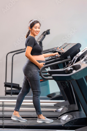 Asian pregnant woman exercising in the gym on treadmill. Fitness and healthy lifestyle during pregnancy and smiling.