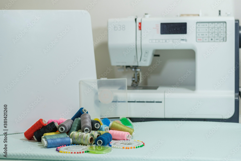 Seamstress's work table - close-up of multi-colored thread coils and other sewing accessories, white electric sewing machine in the background in blur