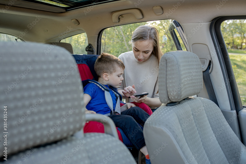 Fototapeta premium Mother gives the phone little boy sitting in a car seat. Safety of transportation of children.