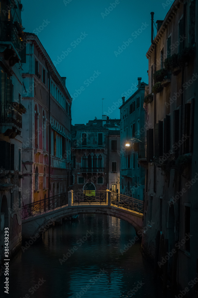 Beautiful small bridge with yellow lighting over a canal in magnificent city of Venice during night time or early evening. Blue hour in Venice, Italy.