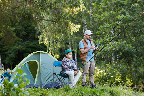 Full length portrait of loving father and son fishing by lake together during camping trip in nature, copy space
