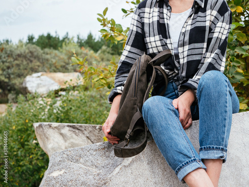 A girl in casual comfortable clothes is sitting on the rocks. A girl in a plaid shirt, blue jeans with a leather backpack, a fashionable autumn look for walking and Hiking. Stylish hipster girl