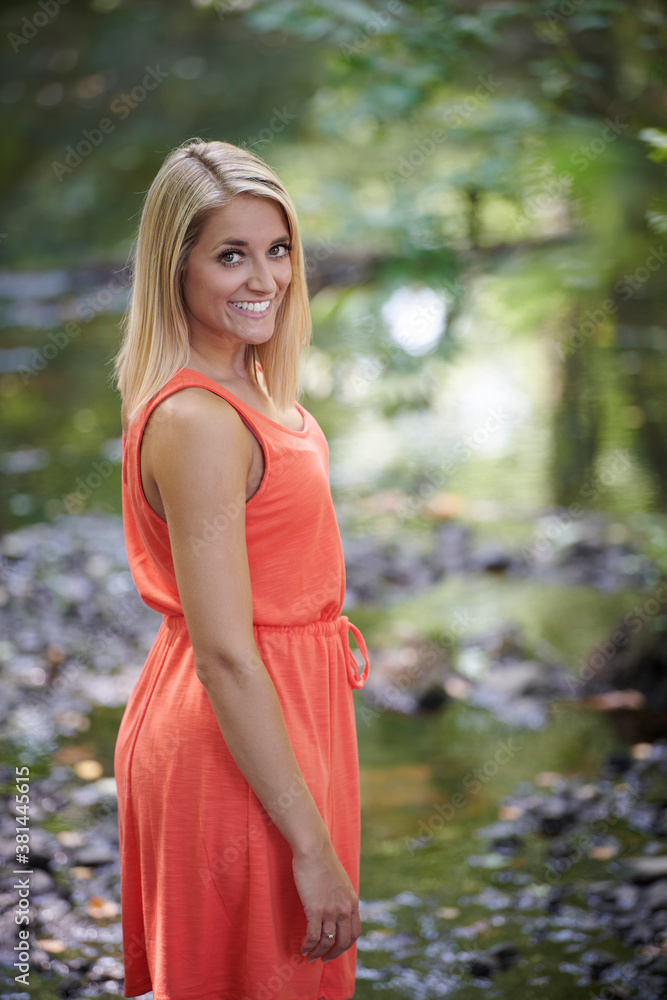 Beautiful young blonde woman poses outside in coral colored sundress stands along small stream in woods Beautiful young blonde woman poses outside in coral colored sundress stands along small stream in woods