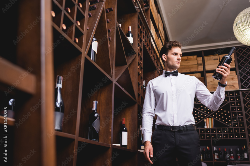 Selective focus of young sommelier in shirt and bow tie holding bottle of wine in restaurant