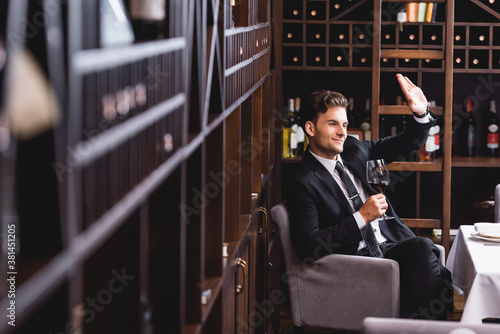 Selective focus of young man in suit waving hand and holding glass of wine in restaurant