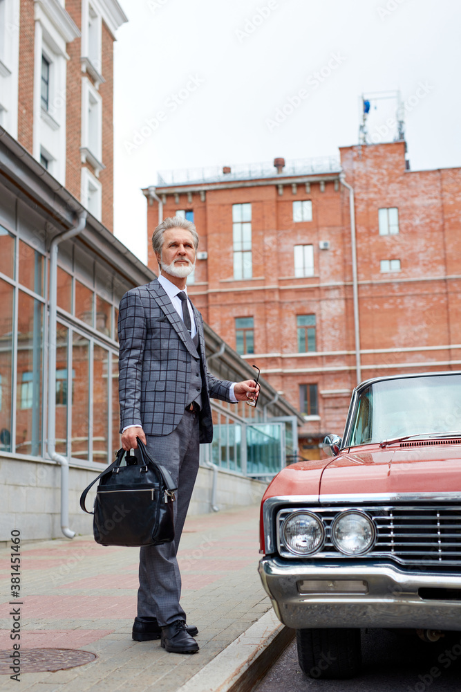 handsome senior businessman near his car outdoors, stylish bearded male in elegant suit after business meeting, next to luxurious representative red auto