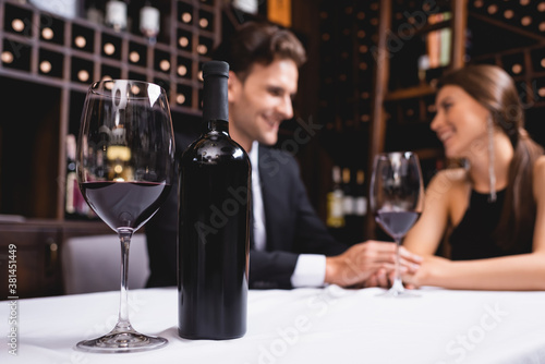 Selective focus of bottle and glass of wine on table near young couple holding hands during dating in restaurant