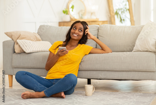 Beautiful black woman with smartphone sitting on floor in living room and studying online from home, copy space
