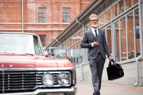 handsome senior businessman near his car outdoors, stylish bearded male in elegant suit after business meeting, next to luxurious representative red auto