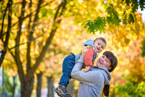 Happy young mother holding sweet toddler boy, family having fun together outside on a nice sunny autumn day. Cute adorable kid and mother play among maple leaves in autumn park or forest