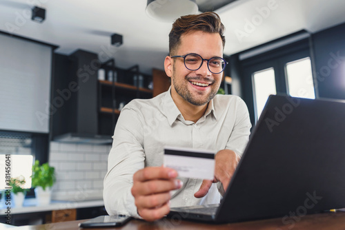 Young man holding credit card sitting in front of laptop computer at home, paying for online order. People, lifestyle, modern technologies and e-commerce concept. Online banking and shopping using mod