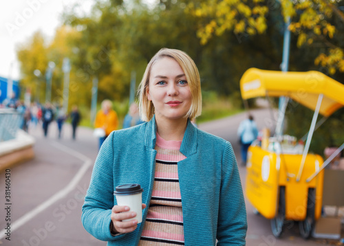 Smiling blonde caucasian woman with coffee cup in autumn park. Outdoor portrait during indian summer.