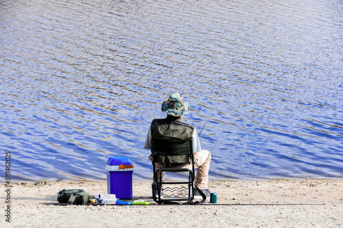 Back foreground of a sitting man angling on the shore of a lake with his spare rod and tackle