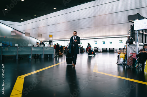 Entrepreneur walking with luggage using mobile phone for checking departure gate at airport terminal, Caucasian corporate director texting via smartphone technology using roaming during business trip