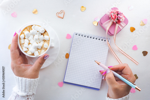 Fotografi female hands with a beautiful manicure write with a pen in a notebook and a white cup with coffee with marshmallows, a pink gift box and heart-shaped sweets