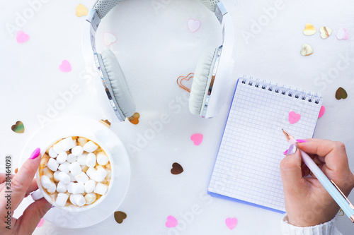 female hands with beautiful manicure hold a cup of coffee and marshmallows and a pen. White headphones with a notepad on a white background with confetti hearts. top view, lifestyle..