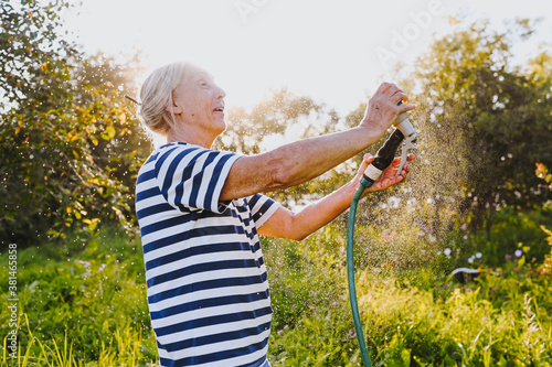 Happy elderly senior woman having fun watering plants with hose in summer garden. Drops of water in backlight. Farming, gardening, agriculture, old age people. Growing organic vegetables on farm