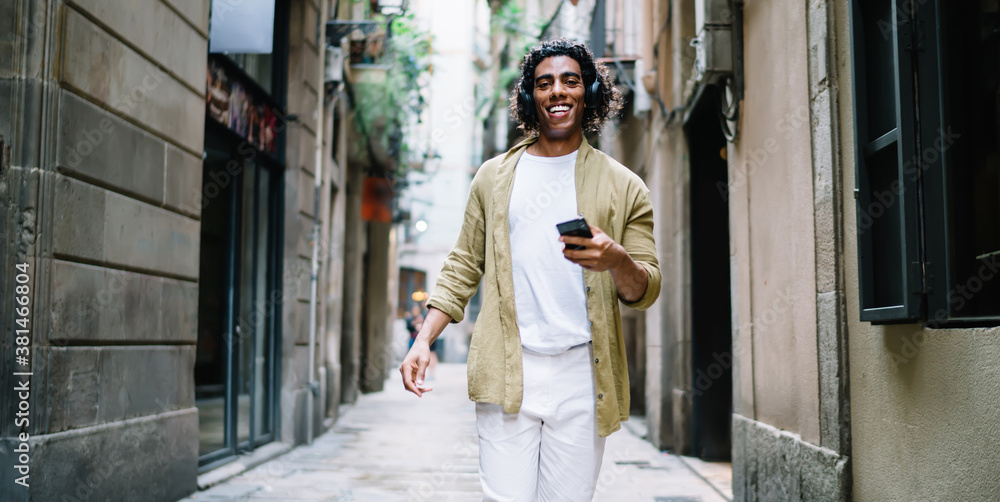 Smiling curly hipster guy in headphones walking on city street listening music and enjoying free time, positive young man meloman holding mobile phone with favorite songs in playlist using earphones