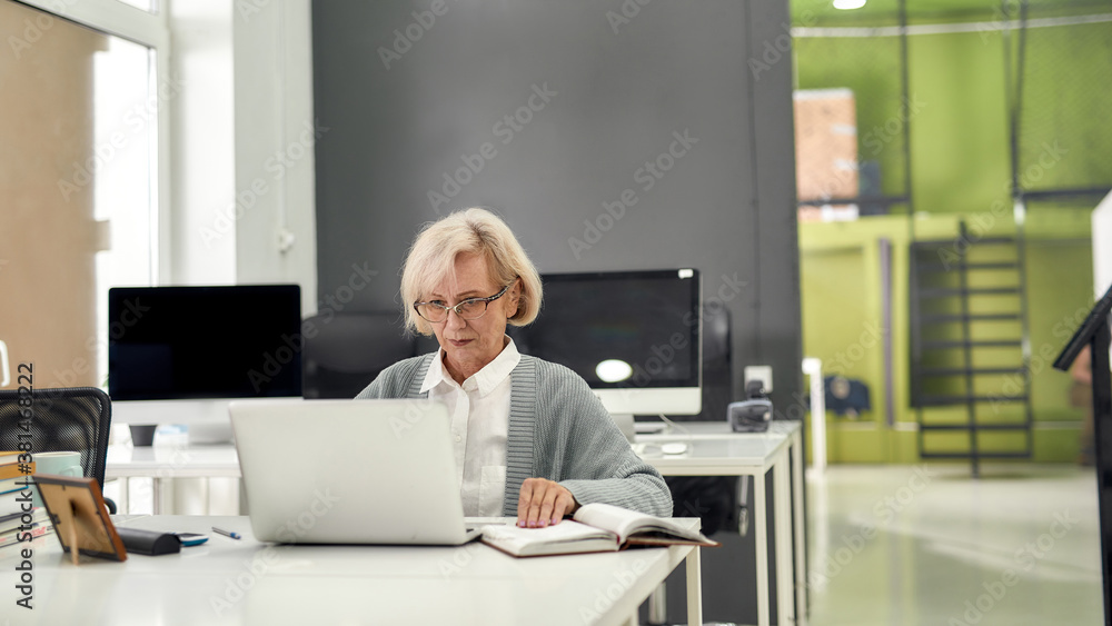 Portrait of aged woman in glasses, senior intern looking focused at the screen while using laptop, sitting at desk, working in modern office