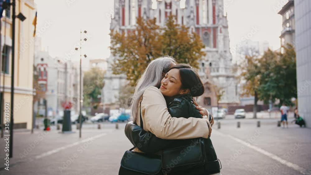 Asian mature mother and adult daughter in casual outerwear are meeting at a city square, smiling, kissing, hugging each other Asian mature mother and adult daughter in casual outerwear are meeting at a city square, smiling, kissing, hugging each other