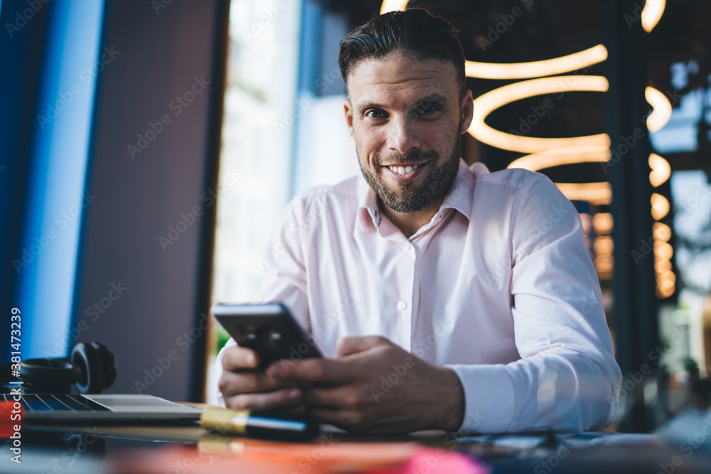 Portrait of millennial Caucasian freelancer smiling at camera enjoying mobile networking via modern cellphone gadget, cheerful successful man with cellular technology posing at table desktop