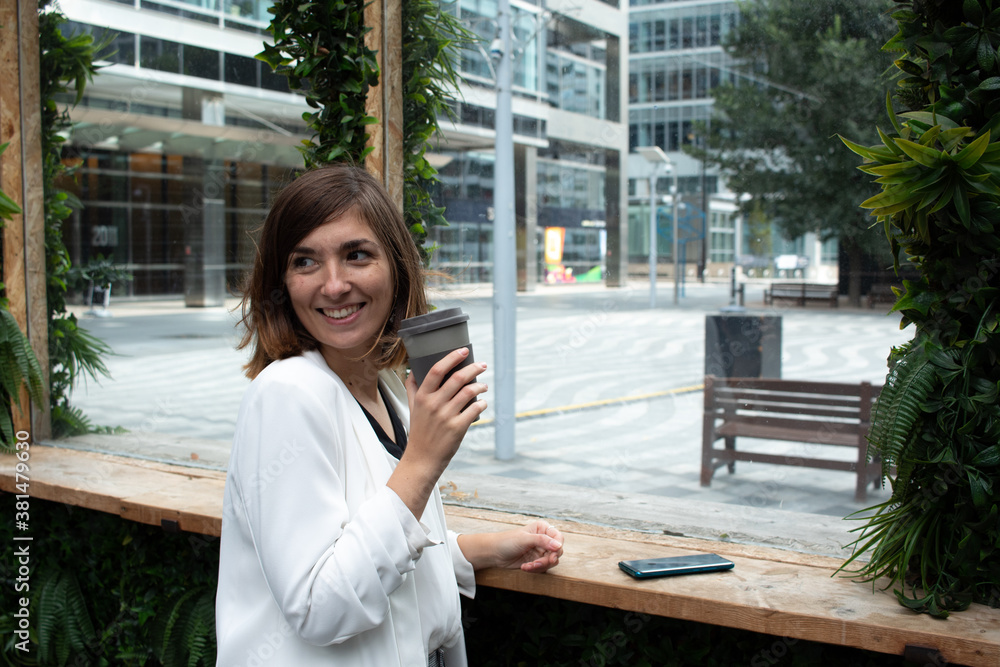 Attractive young female wearing a white suit and drinking coffee from a reusable mug with her smartphone on top of a table in a coffee shop Attractive young female wearing a white suit and drinking coffee from a reusable mug with her smartphone on top of a table in a coffee shop