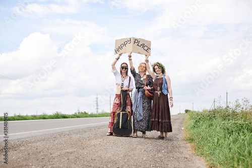 Three hippie women, wearing boho style clothes, standing on road, thumbing a ride, hitchhiking with sign Happy place on cardboard. Friends, traveling together in summer. Freedom and happiness concept.